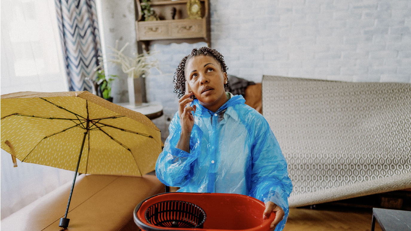 A woman in a waterproof coat, sitting in her home catching flood water in a bucket.