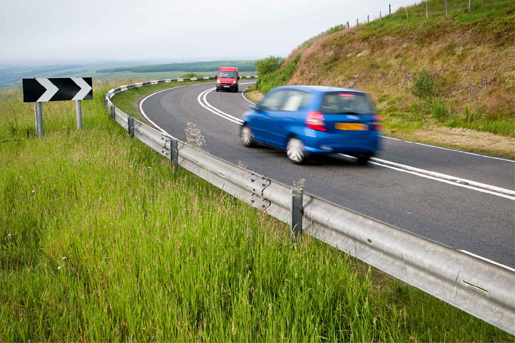 A blue car drives along a winding road, approaching a corner, surrounded by green countryside.