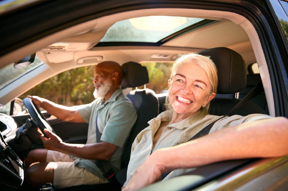 A couple driving in a car in the summer in the UK. A black male is driving, while a white female passenger looks out of her window, smiling